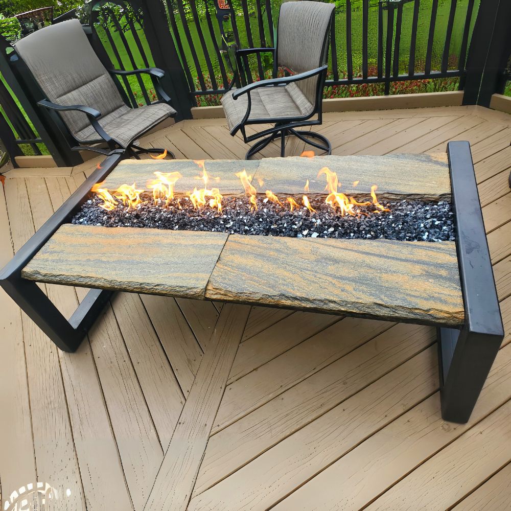 A rectangular Crestone Gas Fire Pit Table with stones on top and visible flames beneath, placed on a deck with outdoor chairs around it.