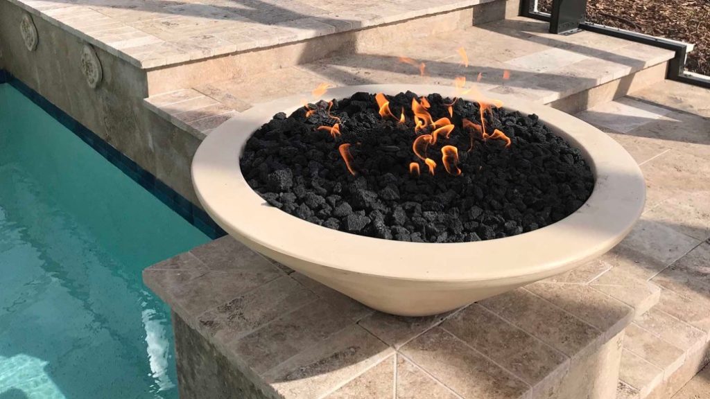 A round, beige concrete gas 48" Cazo Gas Fire Pit with black fire glass inside, placed on a tiled surface next to a swimming pool.