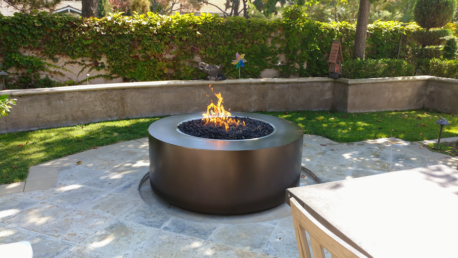 A Unity Round Steel Gas Fire Pit 18" High (3 sizes) with a flame in a backyard patio.